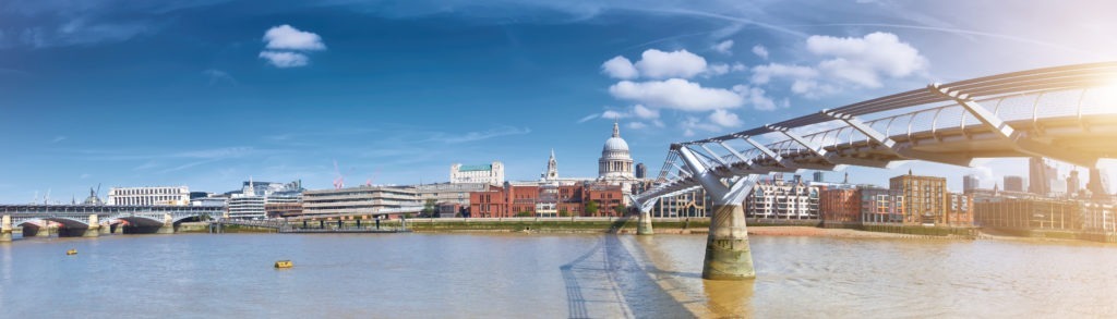 Panoramic view of London featuring the Millennium Bridge stretching across the Thames River. St. Pauls Cathedral, a notable stop on any London Christian Tour, is visible in the background under a clear blue sky. Modern and historic buildings line the riverbank.