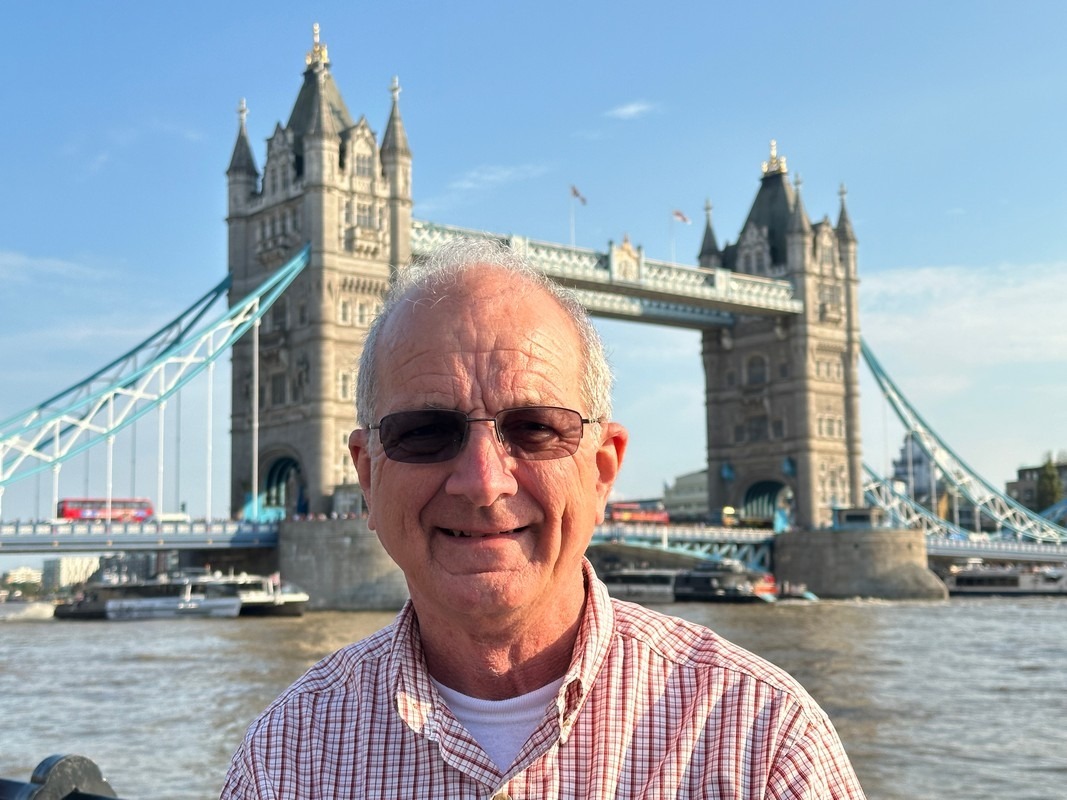 An man wearing sunglasses and a checkered shirt smiles in front of the Tower Bridge, a landmark often admired during London Christian Tours. The historic bridge stands prominently in the background against a clear blue sky.
