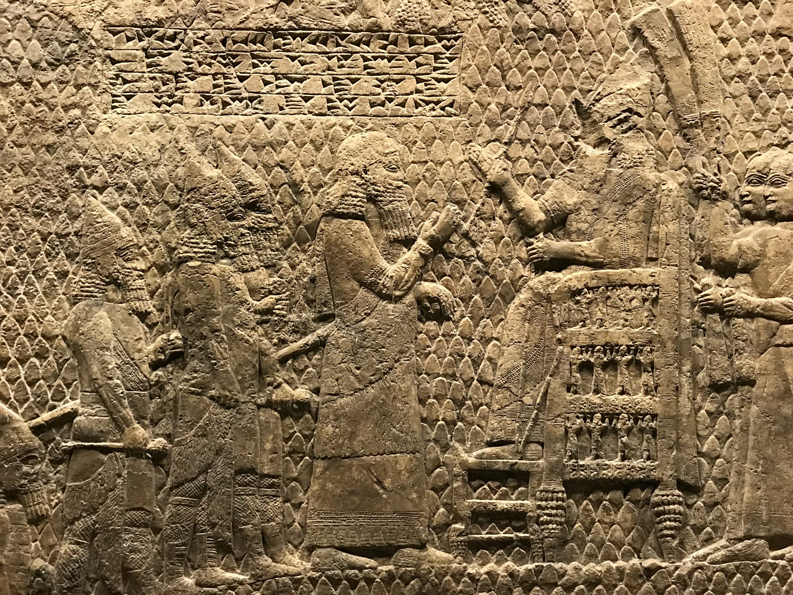 Assyrian Siege of Lachish Sennacherib British Museum Nate Loper. Ancient stone relief depicting figures in elaborate robes; one seated on a throne with others standing before, gesturing as if in conversation. The background features intricate carved patterns. London Christian Tours
